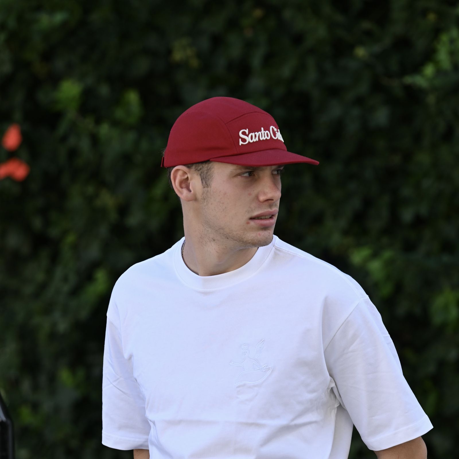 Someone in the red Santo Cielo OG Cap with the embroidered cream script and the white angel tee, in profile against a hedge