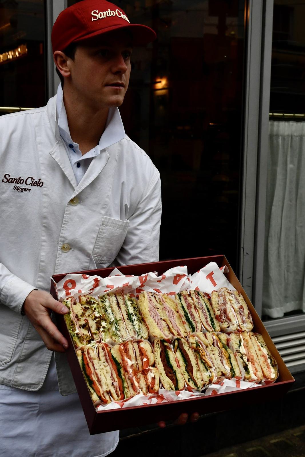 A Santo Cielo baker in whites holding a red platter box of mixed schiacciata sandwiches outside the shop